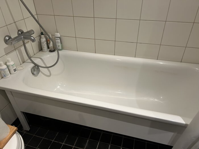 Badkar med duschmunstycke och schampoflaskor i ett kaklat badrum. Lukten misstänks komma från avloppsrör.