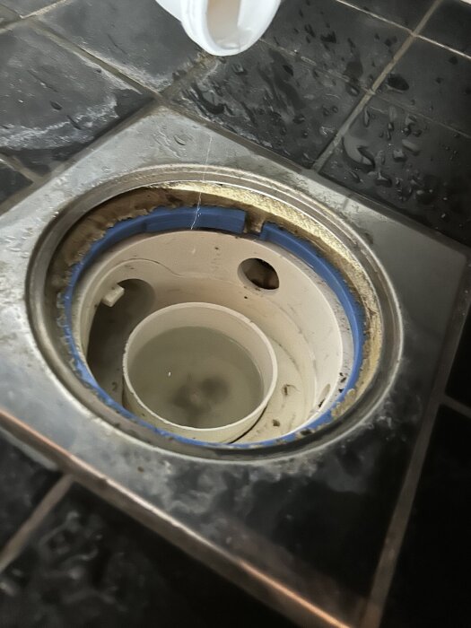 Öppen avloppsbrunn under badkaret med synliga rör och vatten i, omgiven av kakelgolv med vattendroppar.