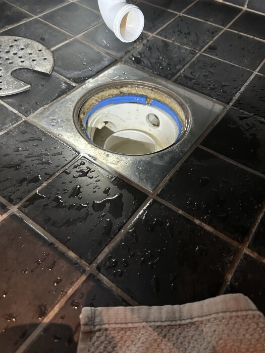 Öppet avloppsgaller på svart kakelgolv i ett badrum, omgiven av vattenstänk och en vit flaska ovanför.