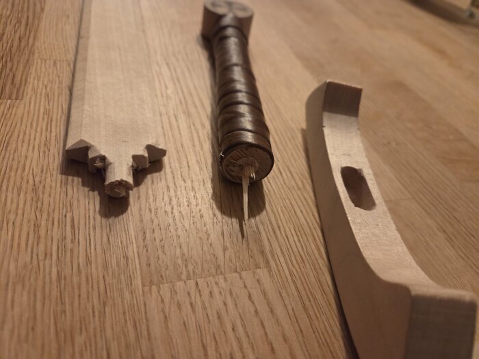 Tre delar av ett trasigt träsvärd på ett trägolv, inklusive blad, handtag med snöre och en sklida med hål.