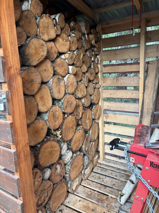Staplade vedträn i ett vedskjul med trä- och nätväggar, under ett tak. En vedklyv står längst bak till höger i bilden.