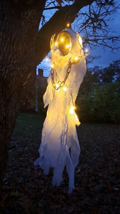 Hängande spökdekoration i ett träd med lysande ljus och kedjor, mot en kvällshimmel.