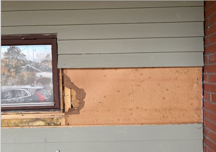 Vägg med grön ytterpanel och ett fönster till vänster. Bakom en skiva syns träfiberskivor och isolering.