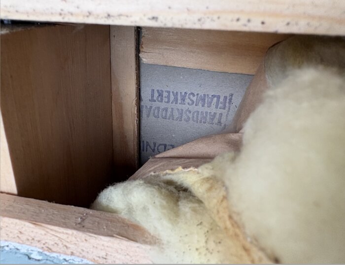 Närbild på träfiberskivor bakom en masonitskiva i en vägg, med synlig glasfiberisolering, del av en renoveringsdiskussion kring isolering.