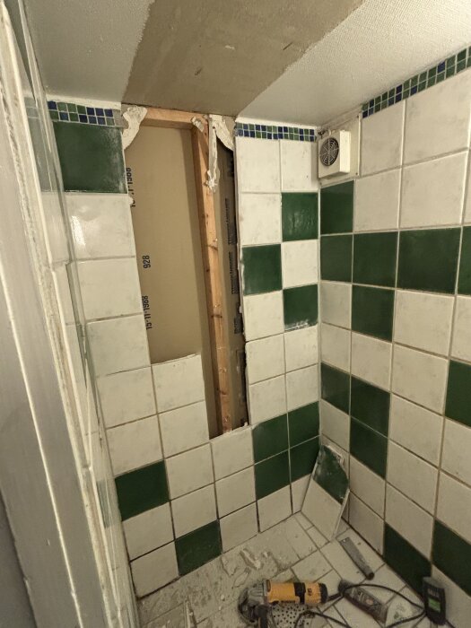 Rivet badrum med gröna och vita kakelplattor, saknar dörr och har verktyg på golvet.