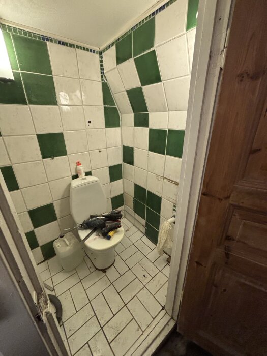 Badrum med gröna och vita golv- och väggplattor; toalettstol fylld med verktyg, och en bensindunk står bredvid. Dörr saknas.