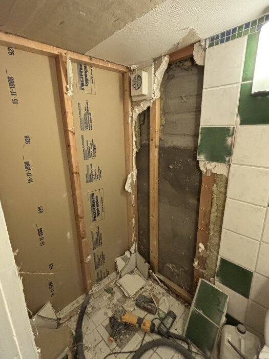 Demontering av badrumsvägg med exponerade träreglar och rivna kakelplattor, verktyg och skräp på golvet.