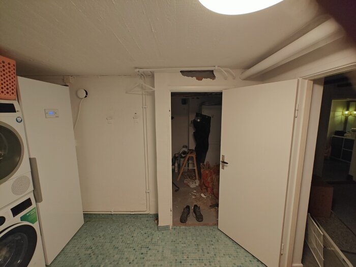 Dörröppning mellan tvättstuga och pannrum med vitmålad vägg; synliga rör i taket och grönt mosaikgolv; arbetsskor och byggmaterial på golvet.