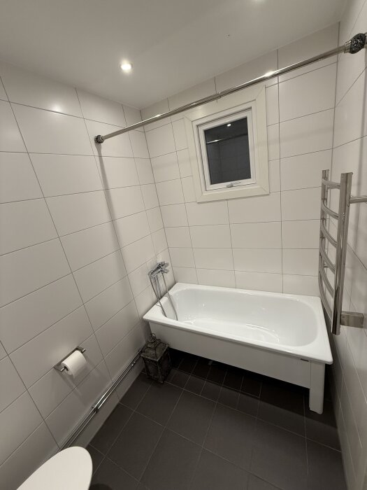 Badrum med ett vitt badkar under ett fönster, vita kakelväggar, och en kromad handdukstork till höger.