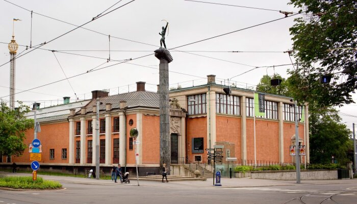 Exteriör av Liljevalchs konsthall i Stockholm, med en pelare och staty i förgrunden samt spårvagnsspår och gatukorsning.