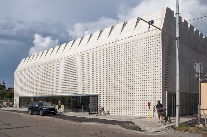 Modernistisk byggnad i betong med perforerad fasad, Liljevalchs konsthall, utan färg eller utsmyckning. Parkerade cyklar och bil utanför ingången.