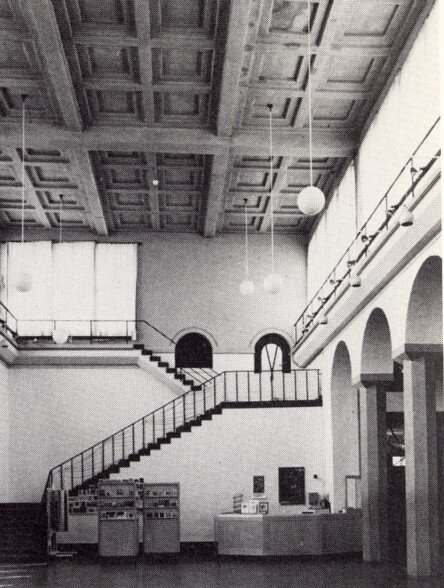 Interiör av byggnad med valv och trappa, dekorativa takdetaljer och hängande lampor, vilket ger en funktionell och minimalistisk estetik.