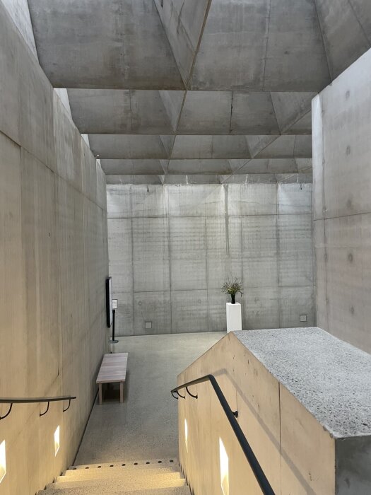 Betonginteriör med minimalistisk design, visar en trappa, bänk och vas på piedestal, kännetecknad av enkla linjer och funktionalitet.