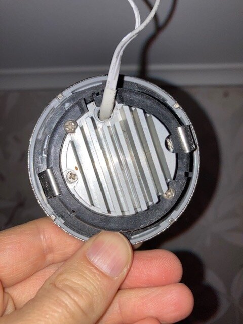 Hand som håller en LED-spotlight med gråmetalliskt hölje och anslutna kablar, fotograferad mot en ljus bakgrund.