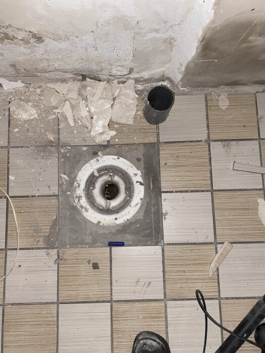 Golvbrunn i källare med kakel runtom, trasigt murbruk och svart avloppsrör. Verktyg och skräp synliga på golvet.