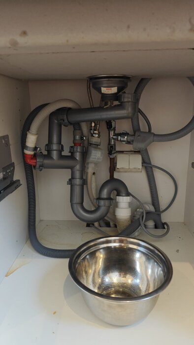 Bild av ett komplicerat avloppssystem under diskbänk med rör och flexibla slangar; en metallskål är placerad under för att fånga upp vatten.