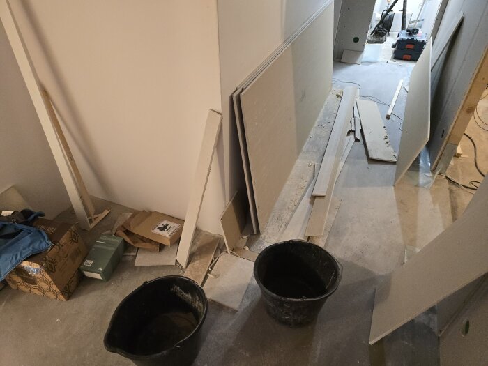 Korridor med byggmaterial och gipsskivor ligger på golvet. Byggplast och verktyg syns i bakgrunden, samt två svarta hinkar i förgrunden.