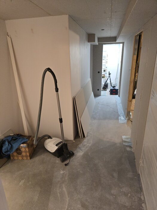 Korridor under renovering med gipsade väggar, dammsugare, kartong och byggmaterial på golvet, kamerautrustning i bakgrunden.