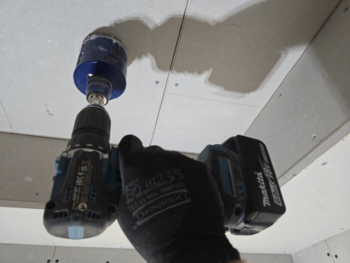 En person använder en batteridriven borrmaskin med en blå hålsåg för att skapa en markering i en gipsvägg.