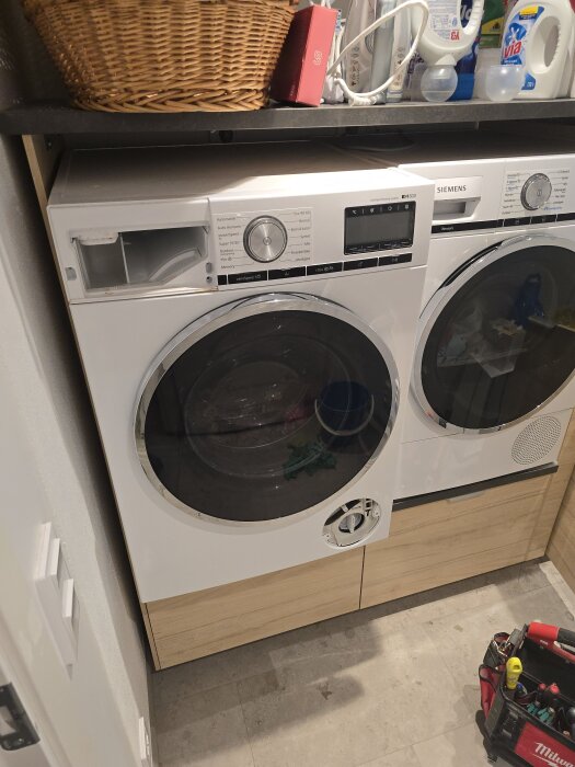 Tvättmaskin och torktumlare i tvättstuga under renovering med verktyg i förgrunden. Siemens toppmodeller, placerade under en hylla med rengöringsmedel.