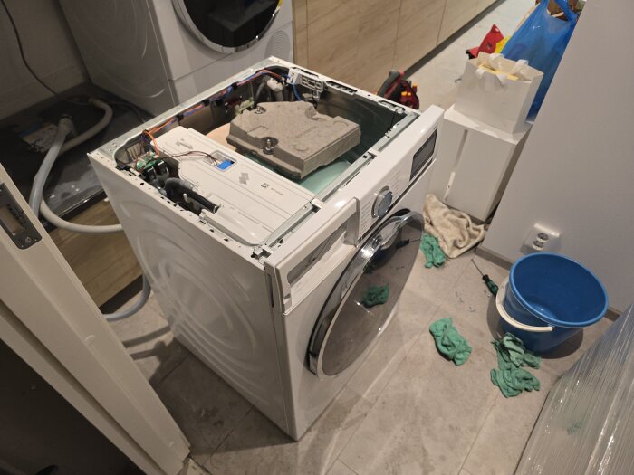 Öppen toppmoderna tvättmaskin under reparation. Verktyg och trasor ligger på golvet i tvättstugan. Blå hink står bredvid maskinen.