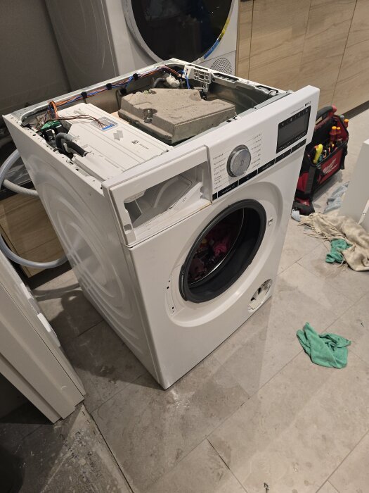 Öppen tvättmaskin under reparation, med avlägsnat toppskydd, elledningar och verktyg i närheten på ett kaklat golv.