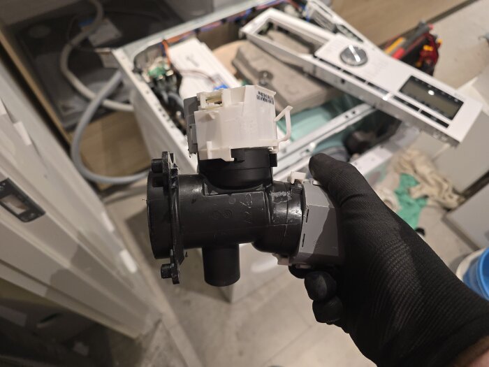 Närbild på en trasig tvättmaskinspump i en handskbeklädd hand, med uppbruten tvättmaskin i bakgrunden.