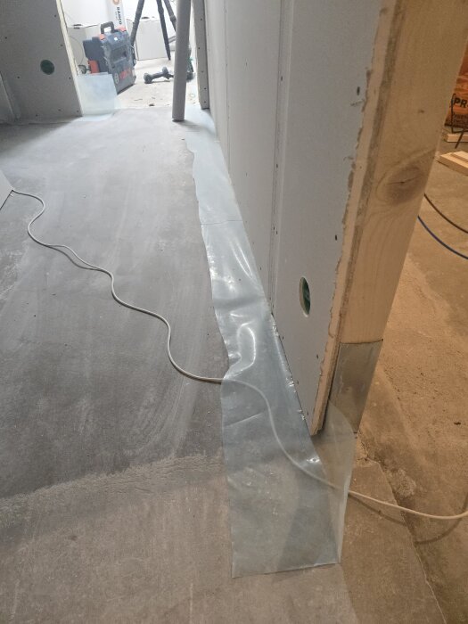 Byggarbetsplats med gipsad vägg och genomskinlig plast längs golvet samt verktyg och utrustning i bakgrunden. En elkabel löper över golvet.