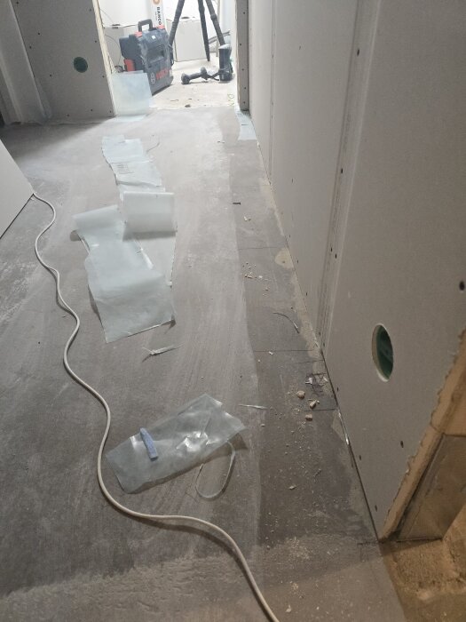 Gipsskivor i en korridor med utspridda plastbitar och en synlig kabel på golvet, verktyg och borrmaskin i bakgrunden.