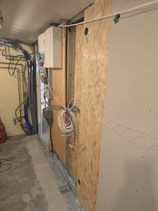 Elektriska ledningar och apparater på en byggvägg med OSB-skiva och gips. Rör, kablar och en elcentral synliga i ett renoveringsprojekt.