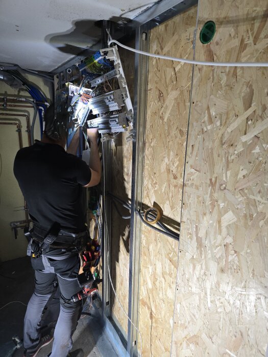 Person arbetar med elcentral och kablar på en plywoodvägg under renovering.