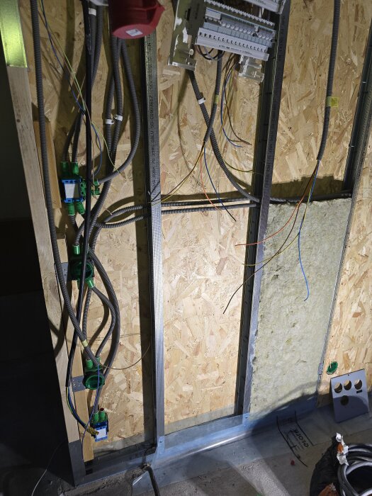Vägg under renovering med målade kablar och elcentraler, isolering och plywood synligt. Kablaget hänger löst genom flexibla rör.