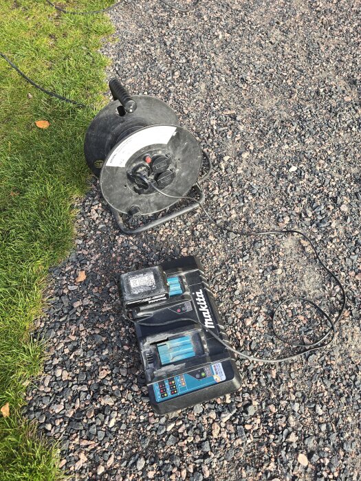 Förlängningskabel och batteriladdare av märket Makita ligger på en grusgång bredvid en gräsmatta.