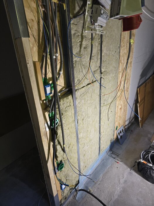 Vägg under renovering med isolering, synliga elledningar och rör. Elcentral hänger löst, omgiven av osb-skivor och gipsskiva ska monteras.