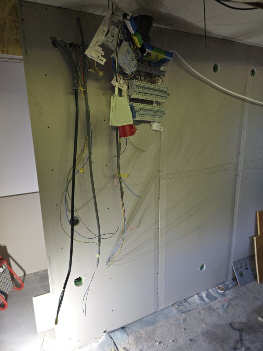 Gipsvägg med synliga kabeldragningar och elcentral under renovering, med hål för elinstallationer och kablar genom väggen.