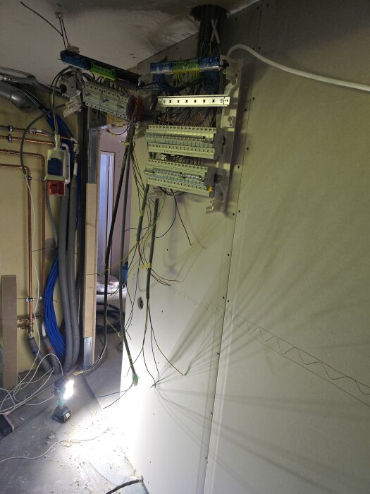 Installation av elektriska kretskort och ledningar bakom en gipsskiva i ett renoveringsprojekt, med blå och gula kablar synliga.