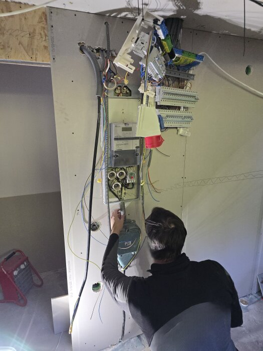 Person installerar elektriska komponenter i ett elskåp på en gipsvägg. Flera kablar hänger löst från väggen.
