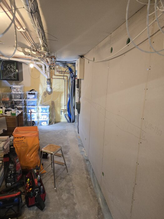 En korridor med gipsväggar, färgmarkerade hål för elinstallation, verktyg och material på golvet, tak med ledningar och rör samt en stege.