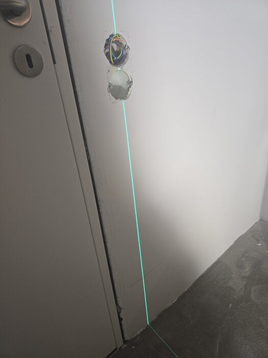 Två borrade hål i en vägg för elinstallation, med synliga kablar och en ljus projicerad vertikal laserlinje som riktmärke.