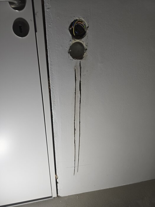 Vägg med förberett uttag och två frästa spår för elektriska ledningar, nära en dörr med synligt dörrlås.