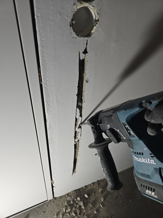 En vägg med ett uthugget spår för elinstallation, ett runt hål upptill och en borrmaskin som används för arbetet.