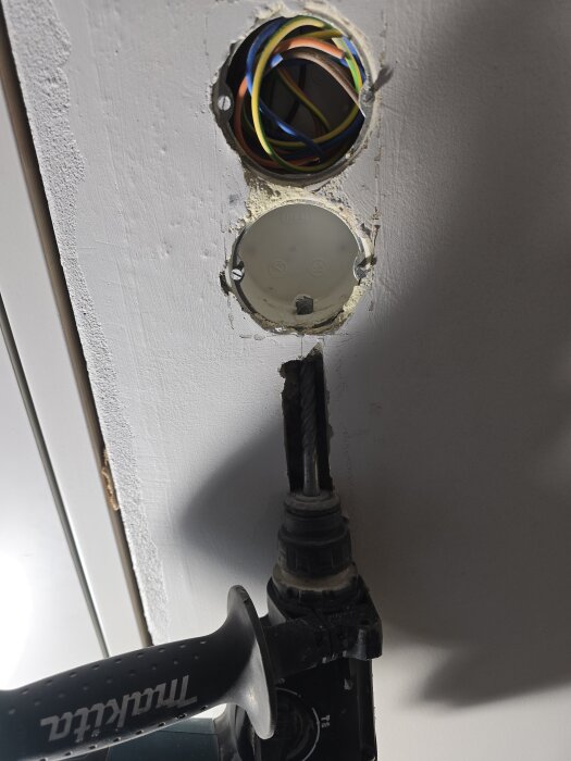 Borrhål i vägg med blottade elkablar, en vit dosa och en borrmaskin under, förberett för installation av vägguttag.