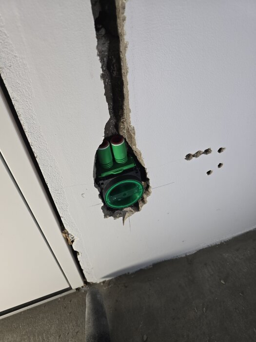 Bilden visar en vägg med ett urfräst hål där en grön el-dosa med anslutningar är installerad, redo för en vägguttag.
