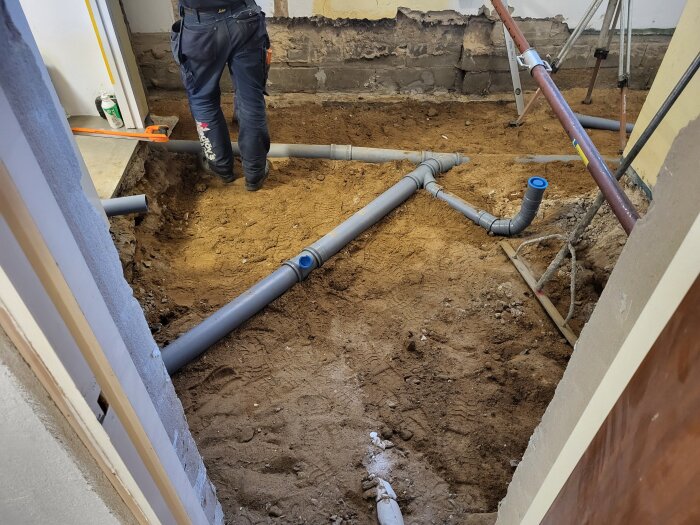 Person arbetar i källare med avloppsrör installerade i utgrävning, väggar delvis oputsade.