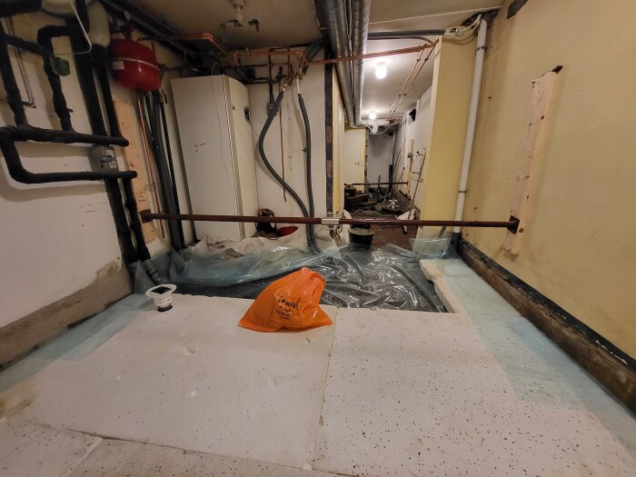Källarrenovering med plastskikt och rörinstallationer, synliga stämpar för stöd, byggmaterial och verktyg i bakgrunden.