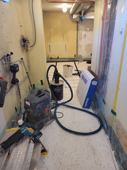 Renovering av källare med verktyg och byggmaterial, inklusive en dammsugare, sågar och rör. Plasten skyddar väggarna, och rör är synliga i taket.