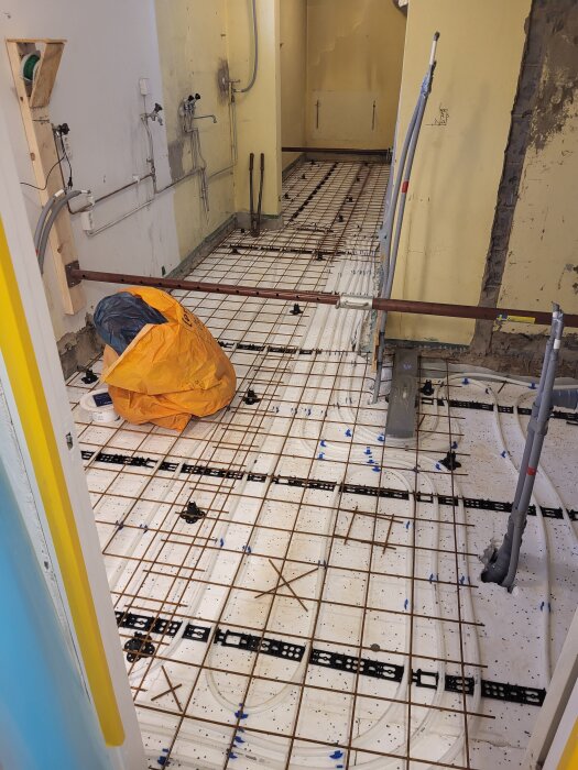 Rum under renovering med exponerad armering och rör dragna längs golvet. Påse med byggmaterial på golvet. Väggarna är gula.