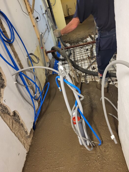 En person häller cement över stålmatta för att göra ett golv. Blå och grå kablar hänger längs väggen.