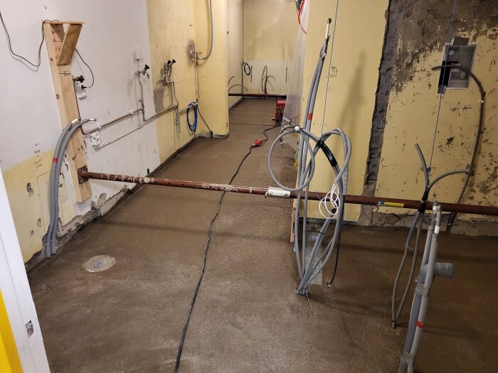 Renoverad källare med nya rör, kablar och gul vägg, saknar ännu ytbehandling. Cementgolv förberett för vidare konstruktion.