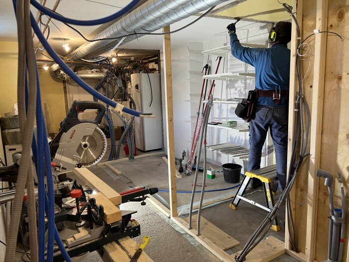 En person arbetar i en renoverad källare med verktyg och ledningar synliga, omgiven av byggmaterial och en snickarbänk.